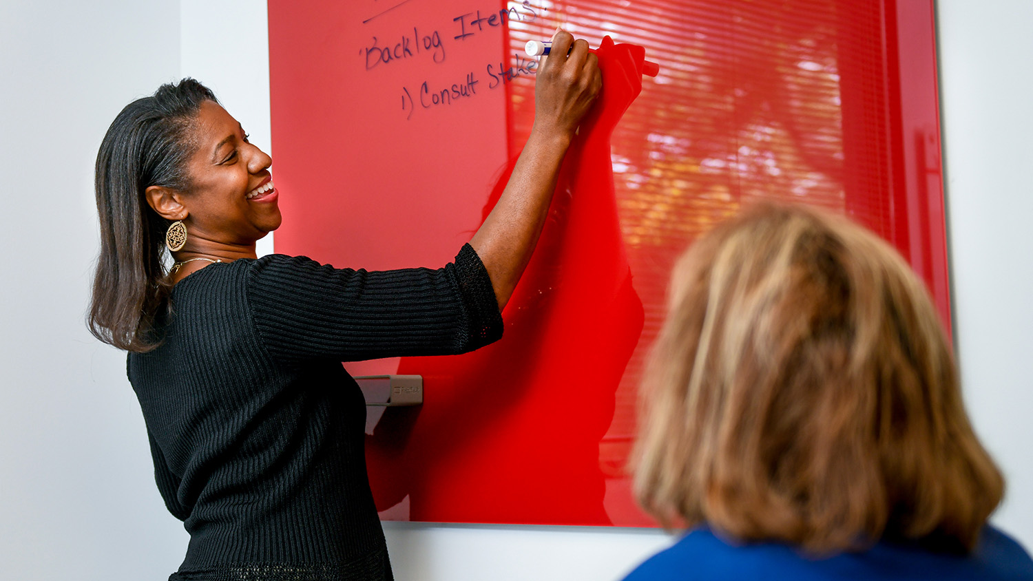 Two University Human Resources employees are in an office. One is sitting, and one is standing at a white board and writing notes.