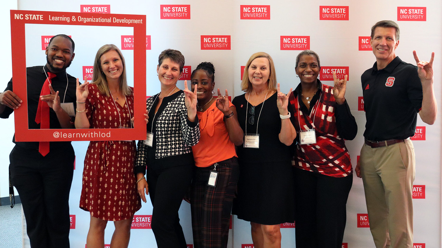 Seven employees pose for a photo in front of a backdrop featuring the NC State brick logo. Two of the employees hold a prop that says "Learning & Organizational Development." All employees make wolf symbols with their hands.